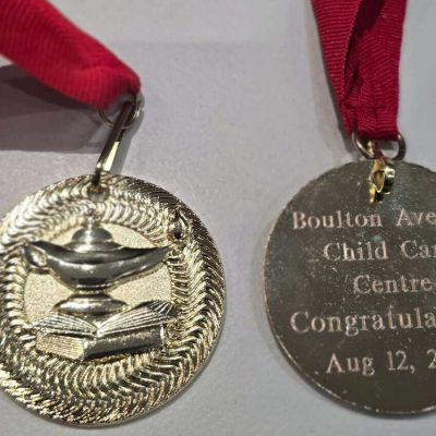 Gold graduation medal with lamp of knowledge and book design, engraved "Boulton Avenue Child Care Centre, Congratulations, Aug 12, 2026," on red ribbon